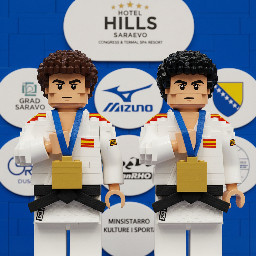 A medium-shot photograph displays two young judo competitors standing side by side against a blue background featuring various sponsor logos. Both wear traditional white judo uniforms, known as *judogis*, secured with black belts. They have sponsor patches on their shoulders, and each wears a large, rectangular gold medal with a blue ribbon around their neck. Both competitors have their right hand resting flat against their chest, over their heart, looking straight ahead with a serious and formal expression.  The competitor on the left has short, curly dark hair, thick eyebrows, and a fair complexion. His judogi has red and yellow stripes on the top of the shoulder and a small rectangular red and yellow patch on the upper right sleeve with the text "ESP". His right hand is placed flat on the left side of his chest. The gold medal hangs from a blue ribbon that has indecipherable white text printed on it.  The competitor on the right also has dark, curly, and slightly longer hair, with thick eyebrows and a fair complexion. His judogi similarly features red and yellow stripes on the shoulders, an "ESP" patch on the right sleeve, and a small circular red and yellow logo on the left side of his chest. His right hand is placed across his chest, in a similar gesture to the first competitor. He wears a gold medal identical to the other athlete's.  The background consists of rounded white panels set against a blue wall. The panels display various commercial and event logos, including "HOTEL HILLS SARAJEVO CONGRESS & TERMAL SPA RESORT" with five stars above, "GRAD SARAJEVO", "Mizuno", "DanRHO", and a logo that includes stars and a blue and yellow shield resembling national symbols of Bosnia and Herzegovina. The words "MINISTARSTVO KULTURE I SPORTA" are also partially visible on one of the panels. The background is slightly out of focus, keeping the two athletes in the foreground as the clear focal point of the image. The lighting is bright and even, typical of an indoor sporting event.