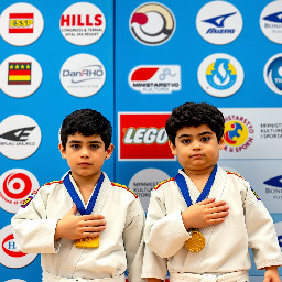 A medium-shot photograph displays two young judo competitors standing side by side against a blue background featuring various sponsor logos. Both wear traditional white judo uniforms, known as *judogis*, secured with black belts. They have sponsor patches on their shoulders, and each wears a large, rectangular gold medal with a blue ribbon around their neck. Both competitors have their right hand resting flat against their chest, over their heart, looking straight ahead with a serious and formal expression.  The competitor on the left has short, curly dark hair, thick eyebrows, and a fair complexion. His judogi has red and yellow stripes on the top of the shoulder and a small rectangular red and yellow patch on the upper right sleeve with the text "ESP". His right hand is placed flat on the left side of his chest. The gold medal hangs from a blue ribbon that has indecipherable white text printed on it.  The competitor on the right also has dark, curly, and slightly longer hair, with thick eyebrows and a fair complexion. His judogi similarly features red and yellow stripes on the shoulders, an "ESP" patch on the right sleeve, and a small circular red and yellow logo on the left side of his chest. His right hand is placed across his chest, in a similar gesture to the first competitor. He wears a gold medal identical to the other athlete's.  The background consists of rounded white panels set against a blue wall. The panels display various commercial and event logos, including "HOTEL HILLS SARAJEVO CONGRESS & TERMAL SPA RESORT" with five stars above, "GRAD SARAJEVO", "Mizuno", "DanRHO", and a logo that includes stars and a blue and yellow shield resembling national symbols of Bosnia and Herzegovina. The words "MINISTARSTVO KULTURE I SPORTA" are also partially visible on one of the panels. The background is slightly out of focus, keeping the two athletes in the foreground as the clear focal point of the image. The lighting is bright and even, typical of an indoor sporting event.