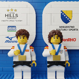 A medium-shot photograph displays two young judo competitors standing side by side against a blue background featuring various sponsor logos. Both wear traditional white judo uniforms, known as *judogis*, secured with black belts. They have sponsor patches on their shoulders, and each wears a large, rectangular gold medal with a blue ribbon around their neck. Both competitors have their right hand resting flat against their chest, over their heart, looking straight ahead with a serious and formal expression.  The competitor on the left has short, curly dark hair, thick eyebrows, and a fair complexion. His judogi has red and yellow stripes on the top of the shoulder and a small rectangular red and yellow patch on the upper right sleeve with the text "ESP". His right hand is placed flat on the left side of his chest. The gold medal hangs from a blue ribbon that has indecipherable white text printed on it.  The competitor on the right also has dark, curly, and slightly longer hair, with thick eyebrows and a fair complexion. His judogi similarly features red and yellow stripes on the shoulders, an "ESP" patch on the right sleeve, and a small circular red and yellow logo on the left side of his chest. His right hand is placed across his chest, in a similar gesture to the first competitor. He wears a gold medal identical to the other athlete's.  The background consists of rounded white panels set against a blue wall. The panels display various commercial and event logos, including "HOTEL HILLS SARAJEVO CONGRESS & TERMAL SPA RESORT" with five stars above, "GRAD SARAJEVO", "Mizuno", "DanRHO", and a logo that includes stars and a blue and yellow shield resembling national symbols of Bosnia and Herzegovina. The words "MINISTARSTVO KULTURE I SPORTA" are also partially visible on one of the panels. The background is slightly out of focus, keeping the two athletes in the foreground as the clear focal point of the image. The lighting is bright and even, typical of an indoor sporting event.